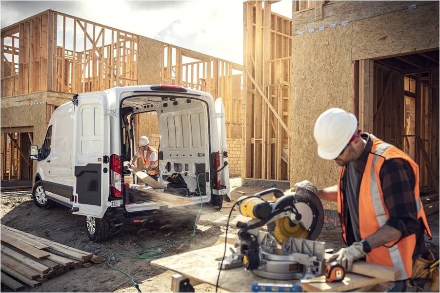 A white Ford E-Transit van with a man outside using an electric saw that is plugged into the van and a building in construction in the background.