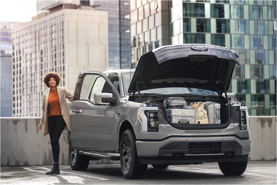 : A gray Ford F-150 Lightning with the front trunk open and a woman standing by an open passenger door.